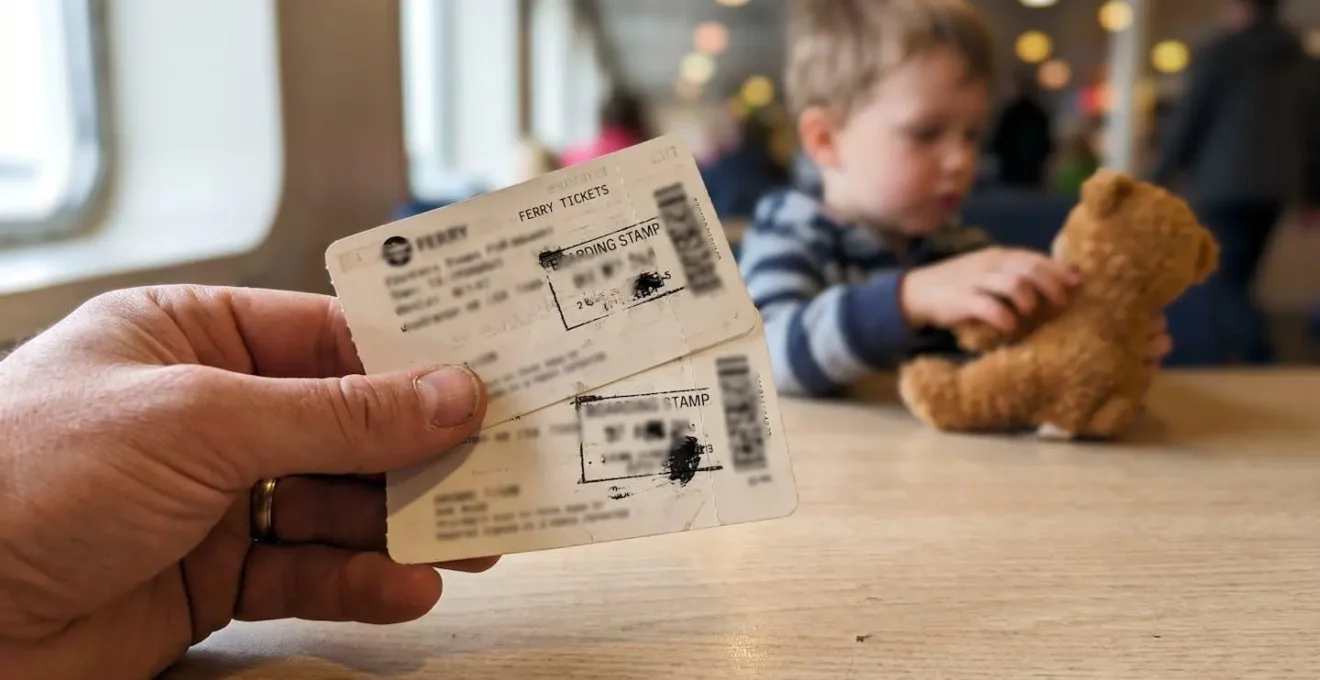 Gros plan sur une main adulte tenant des billets de ferry avec tampons d'embarquement légèrement flous, arrière-plan doux montrant un enfant jouant avec une peluche